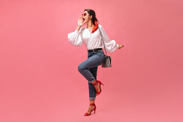 Woman in good mood shouts on pink background. Stylish girl in white blouse, jeans and red heels posing with striped handbag..