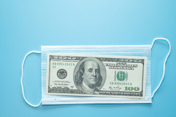 hundred dollars bill lay on medical mask on blue background, concept - cost of life