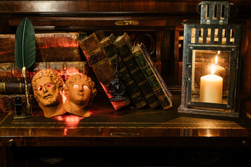 antique books stacked with a pocket watch. They're on a wooden desk on leather. It incorporates two masks of Roman gods and a pen with an inkwell