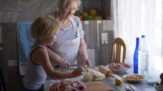 Funny blond boy helps beloved  grandmother in kitchen. elderly woman rolls pastry for pasties and puts minced meat. teenager touches dough. Kid and granny are happy together. shallow depth of field 