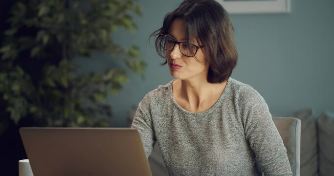 Cheerful dark-haired woman talking and gesturing during video call on portable computer. Beautiful lady using laptop for online conversation while sitting at living room.