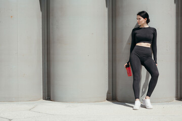 Full-length portrait of a sports woman in a black uniform holding water bottle and looking into distance on a street in a park