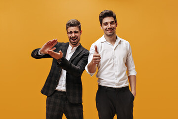 Bearded man in checkered suit claps hands and smiles. Brunet guy in white shirt shows thumbs up. Businessmen pose on orange background.