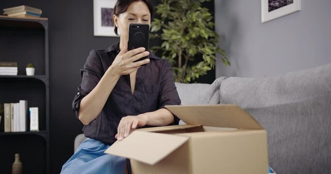 Attractive woman in stylish outfit recording video on smartphone while doing unpacking of delivered parcel. Smiling female blogger making new content for her vlog.