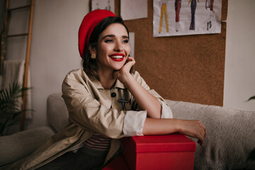 Charming lady in red stylish beret smiling and sitting on sofa. Wonderful young woman with dark hair in beige coat posing..