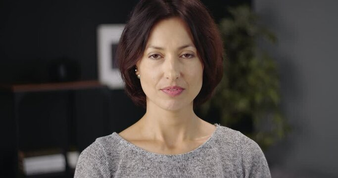 Portrait of attractive woman with dark hair standing in modern apartment and posing on camera. Mature lady in casual outfit smiling and looking ahead.
