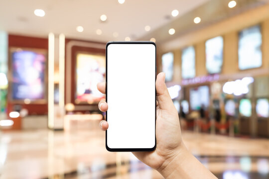 Mockup blank white screen mobile phone hand holding smartphone  with blurred image hall of ticket sales counter at movie theater. Background concept for buy cinema tickets, concerts, etc