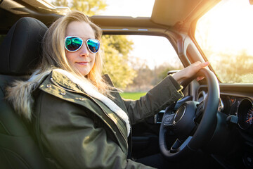 Naklejka premium Blonde female driver, driving in nature in convertible with aviator sunglasses and jacket, during sunset