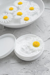 Fototapeta na wymiar Chamomile natural cream. Chamomile in an opened jar with white face and body cream. Plate with chamomile flowers in water on a textured background. Top view.