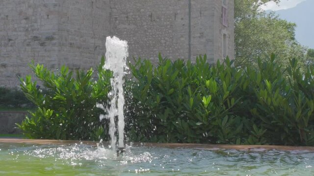 Slowmotion of a small fountain in the center of Riva Del Garda with some green plants and the wall of an old castle in the background
