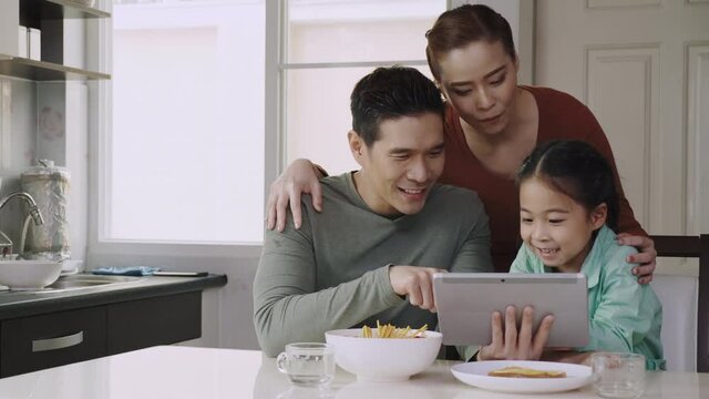 Happy Asian family. Mom and dad are playing a tablet with a cute daughter on the dining table in the home. People are video calling with relative or friend. Using high speed internet technology