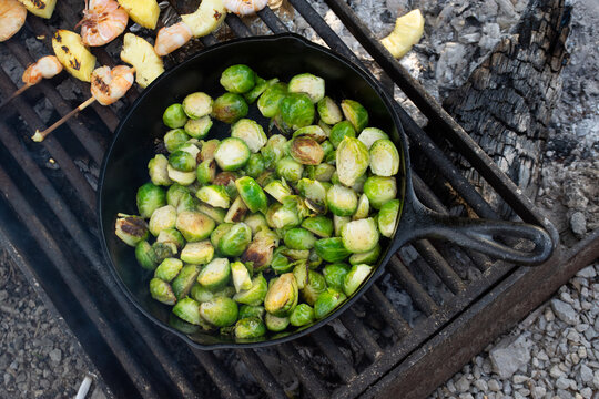 Brussels Sprouts over camp fire in a cast iron skillet with shrimp and pineapple skewers in background