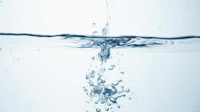 Freshness of a clear blue liquid on white background. Brilliant stream of water in slow motion falls on smooth clean surface, creating air bubbles, drop splashes and ripples after falling, side view