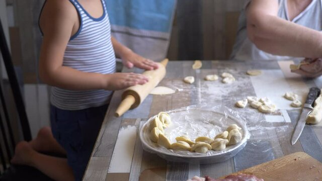 Funny blond boy helps beloved  grandmother in kitchen. elderly woman rolls pastry for pasties and puts minced meat. teenager touches dough. Kid and granny are happy together. shallow depth of field 