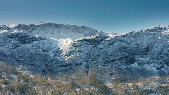 Mountain range rising above the Gaula river valley. Dense pine forest growing on mountainsides and foothills. The thin layer of fresh snow covering the landscape. Blue cloudless skies above.