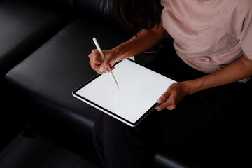 Young woman using black tablet device with white screen. Woman holding tablet, scrolling pages while sitting on the sofa in the living room.