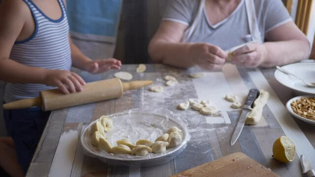 Funny blond boy helps beloved  grandmother in kitchen. elderly woman rolls pastry for pasties and puts minced meat. teenager touches dough. Kid and granny are happy together. shallow depth of field 