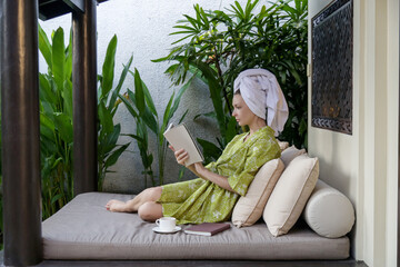 Obraz premium Young caucasian woman in robe and hair wrapped in towel sitting at home and reading a book. 