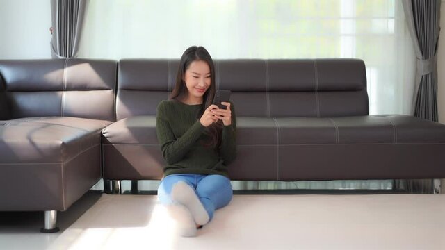 Pretty Asian Female Looking at Smartphone and Smiling While Sitting on Floor by Sofa in Family Home