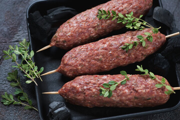 Close-up of raw beef lula kebabs in a cast-iron tray with fresh thyme and charcoals, selective focus
