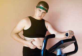 Cheerful fat man in a funny tracksuit is engaged in a stationary bike. The concept of problems with...