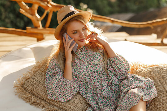 Young beautiful girl in straw hat against lake in city park speaks by mobile phone. Happy blond torist woman in straw use phone