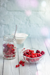 Currants and cherries in a glass jar, a glass plate with berries and sugar on the background of a table of white boards; light background for summer recipes for making homemade jam