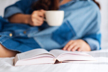 A beautiful young woman reading book and drinking hot coffee in a white cozy bed at home