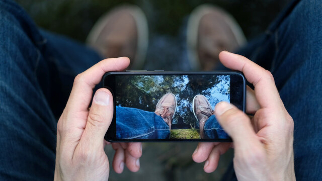 POV men taking feet picture with smartphone sitting on old bridge in park. Top view male legs dangling over water. Guy making photos for social network. Personal perspective, relaxing and mindfulness.