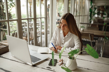 Gorgeous Caucasian hipster young woman in glasses plans her day in a notebook touching his head and...