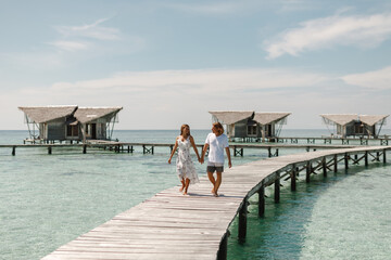 Happy loving couple walking on summer tropical beach on wooden jetty with overwater wooden bungalow on background