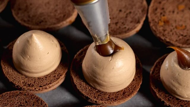Chef is making caramel macarons. Hand with confectionery bag squeezing caramel in cream filling, close-up.