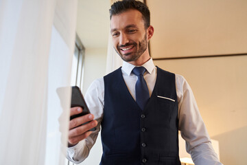 Happy young bearded executive using smartphone indoors
