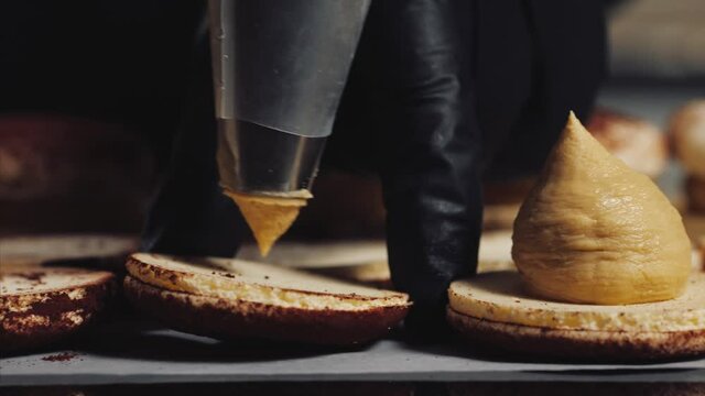 Chef is making caramel macarons. Hand with confectionery bag squeezing cream filling to macarons shells, close-up.