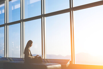 Silhouette of young asian woman is sitting in departure terminal of the airport building, while is using digital tablet for connect to wireless.Asian female entrepreneur is sitting front office window