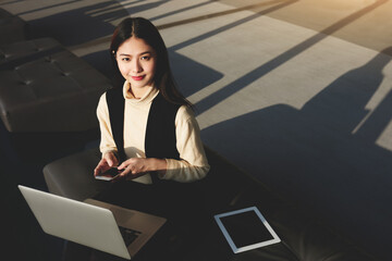 Asian female managing director is using mobile phone and portable laptop computer, while is waiting in airport hall for international partners. Young woman with cellphone in hands is looking in camera