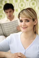 Woman smiling at camera, man reading newspaper in the background