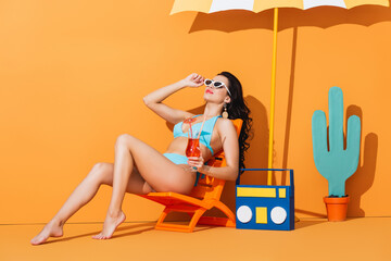 stylish girl in sunglasses and swimsuit sitting on deck chair near paper boombox, cactus and...