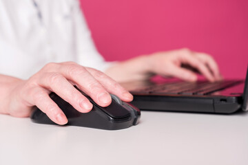 Close up f an elderly woman hands on mouse and laptop keyboard. She plays computer games or searching in web, chatting in social media sites. White table and pink background.