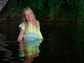 Fototapeta premium blonde girls with blue eyes bathes in clothes in the dark water of a lake at sunset