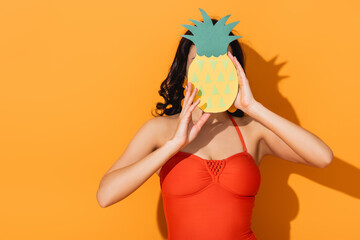 woman in swimsuit covering face with paper cut pineapple on orange