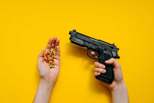 Human hand holding a plastic toy gun and a bunch of plastic ball bullets on bright clean yellow background.