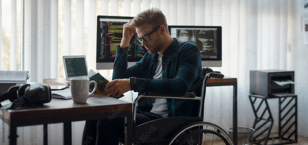 Wall mural more knowledge. portrait of concentrated male web developer in a wheelchair reading a book while sit