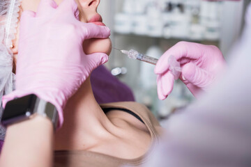 Cosmetologist doctor do injection procedure to a woman in her clinic. She wear glowes and mask and holds syringe with a needle