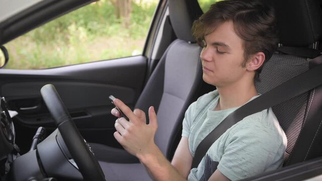 Teen boy on driver seat scrolling smartphone in car parked next to road.