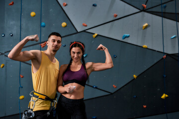 Young male instructor and woman looking at camera, showing biceps, standing against artificial...