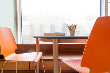 Desk with book and pencils at window. 2 orange chairs.