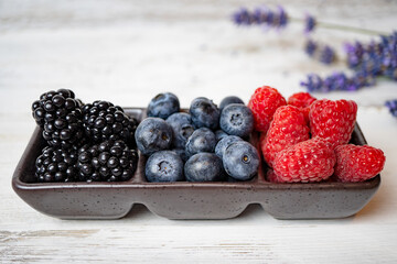 A plate with tasty summer juicy berries (raspberries, blackberries, blueberries) stands on a white wooden background. Nearby are lavender flowers. Fresh seasonal berries. Vegetarian, detox food.