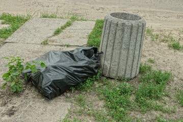 one gray concrete urn and a black plastic bag with garbage on the ground and green grass on the street