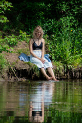 A girl with blond hair on the lake shore has her feet in the water and is swinging them creating a spray of water. Girl in a denim suit in nature